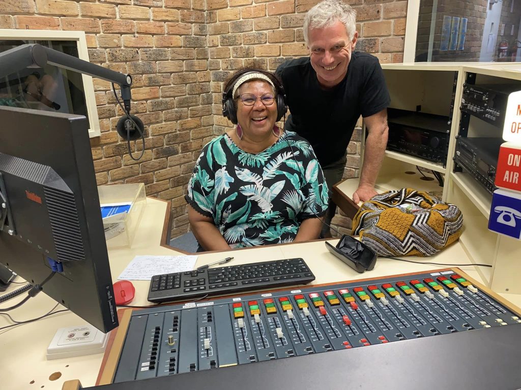 Broadcaster Maureen Mopio-Jane, a woman wearing headphones and a printed top, and Stefan Armbruster, a white man in a black T-shirt, behind a radio production desk. They are both smiling.